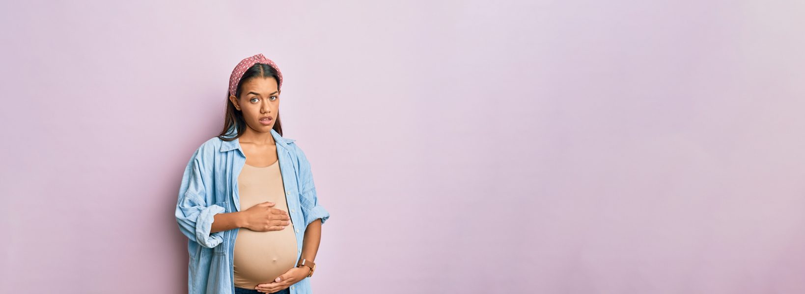 pregnant woman in a tan shirt and chambray jacket in front of a light purple background